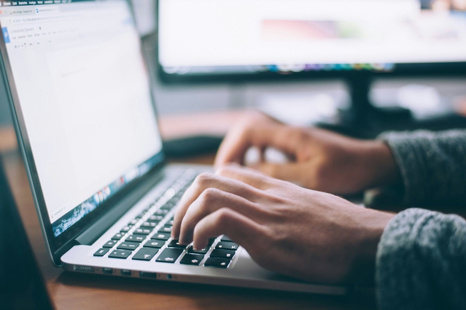 A close-up view of someone's hands typing on a laptop. In the background you can see their second screen.