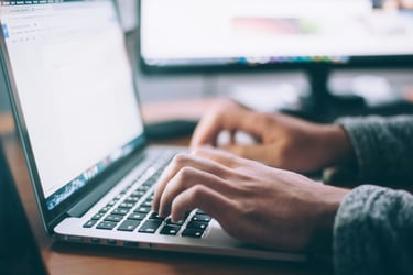 A close-up view of someone's hands typing on a laptop. In the background you can see their second screen.
