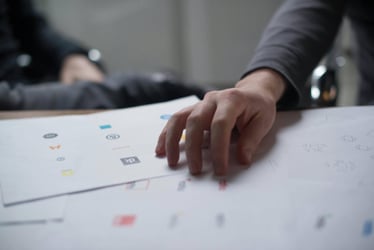 Printed sheets sit on a desk covered in logo designs. A hand rests on top as two people review the designs.